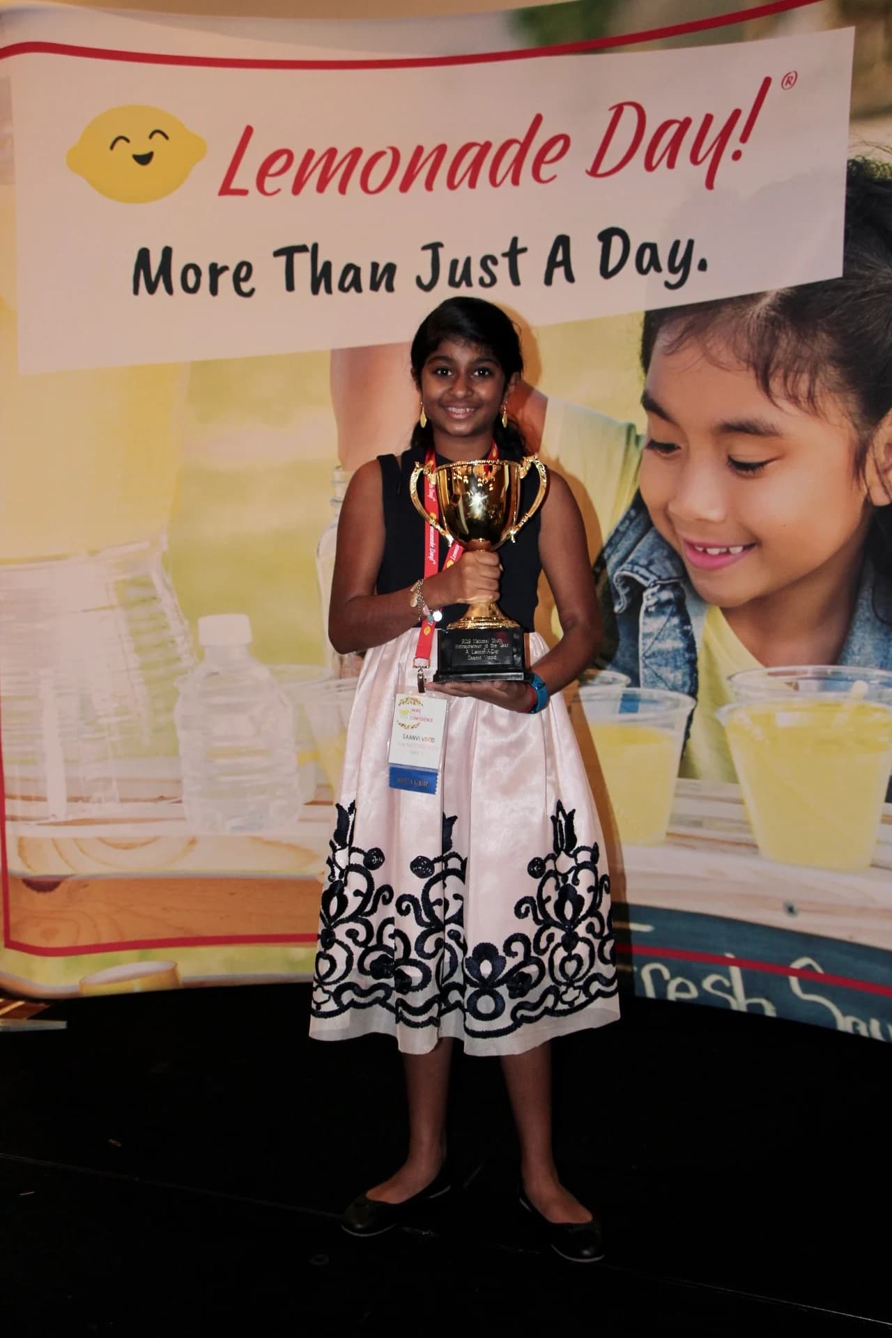 Saanvi holding her trophy in front of a Lemonade Day banner