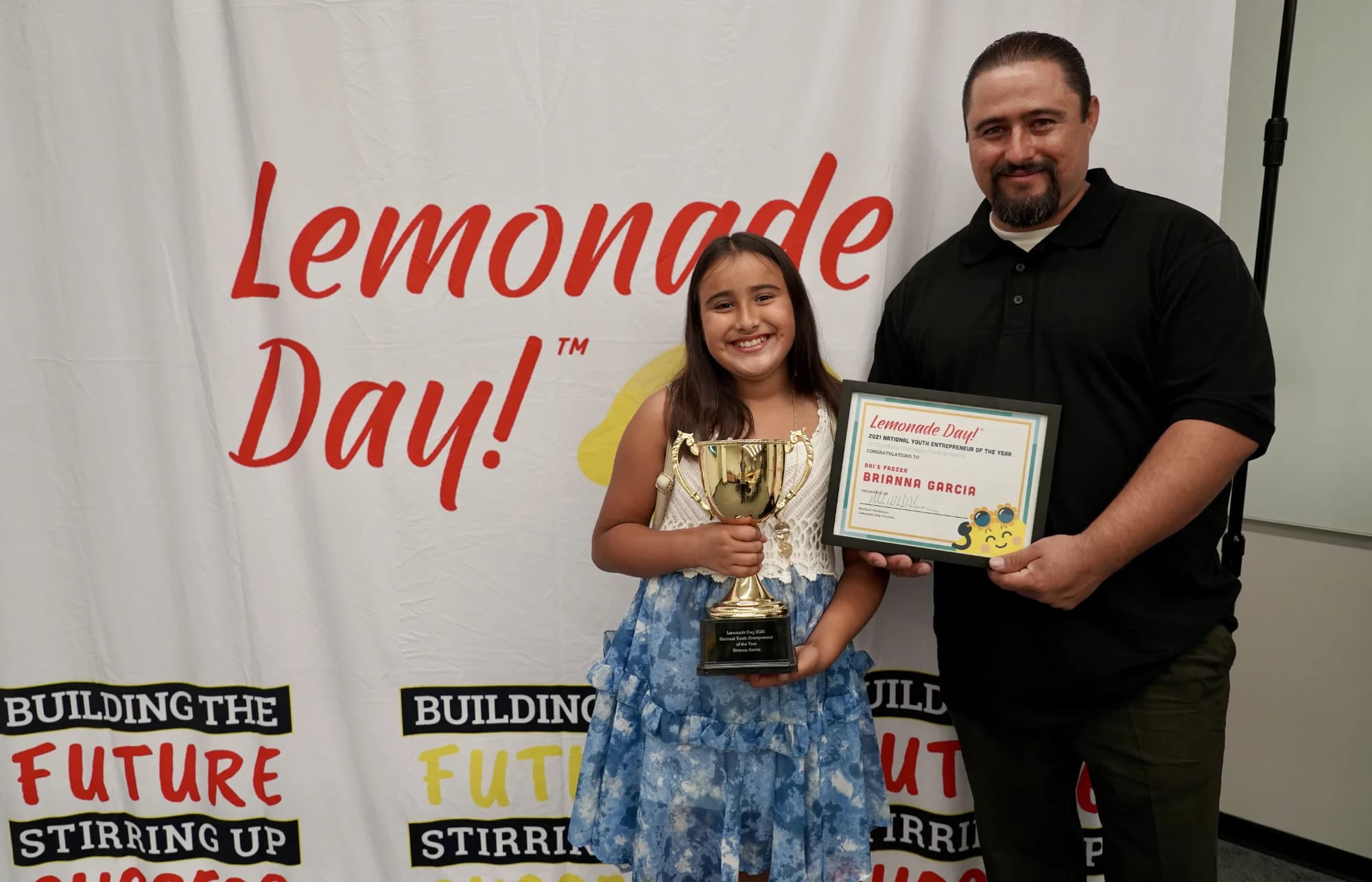 Brianna and her dad holding her award in front of the Lemonade Day backdrop