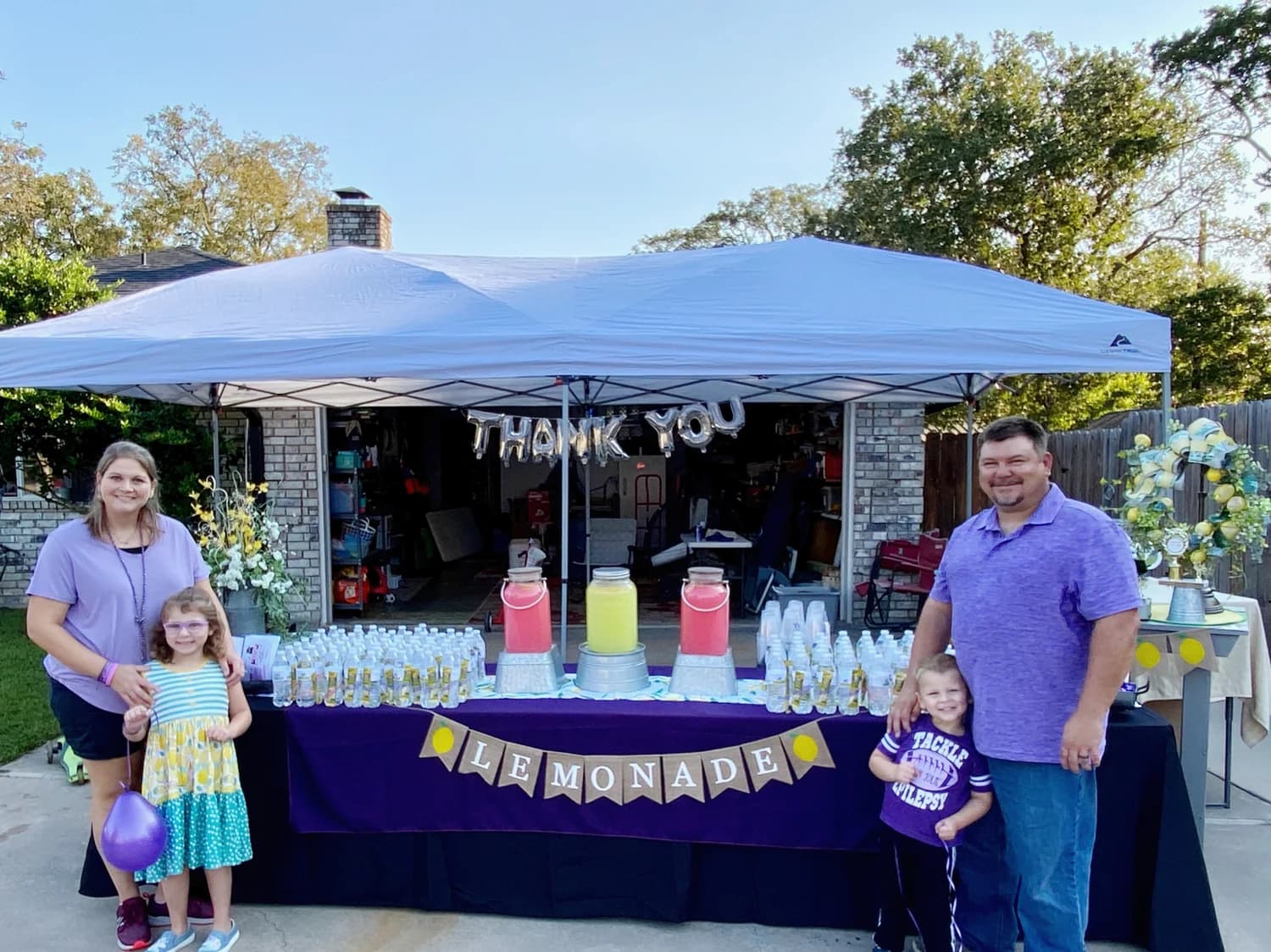 A family lemonade stand under a blue tent with a Thank You banner