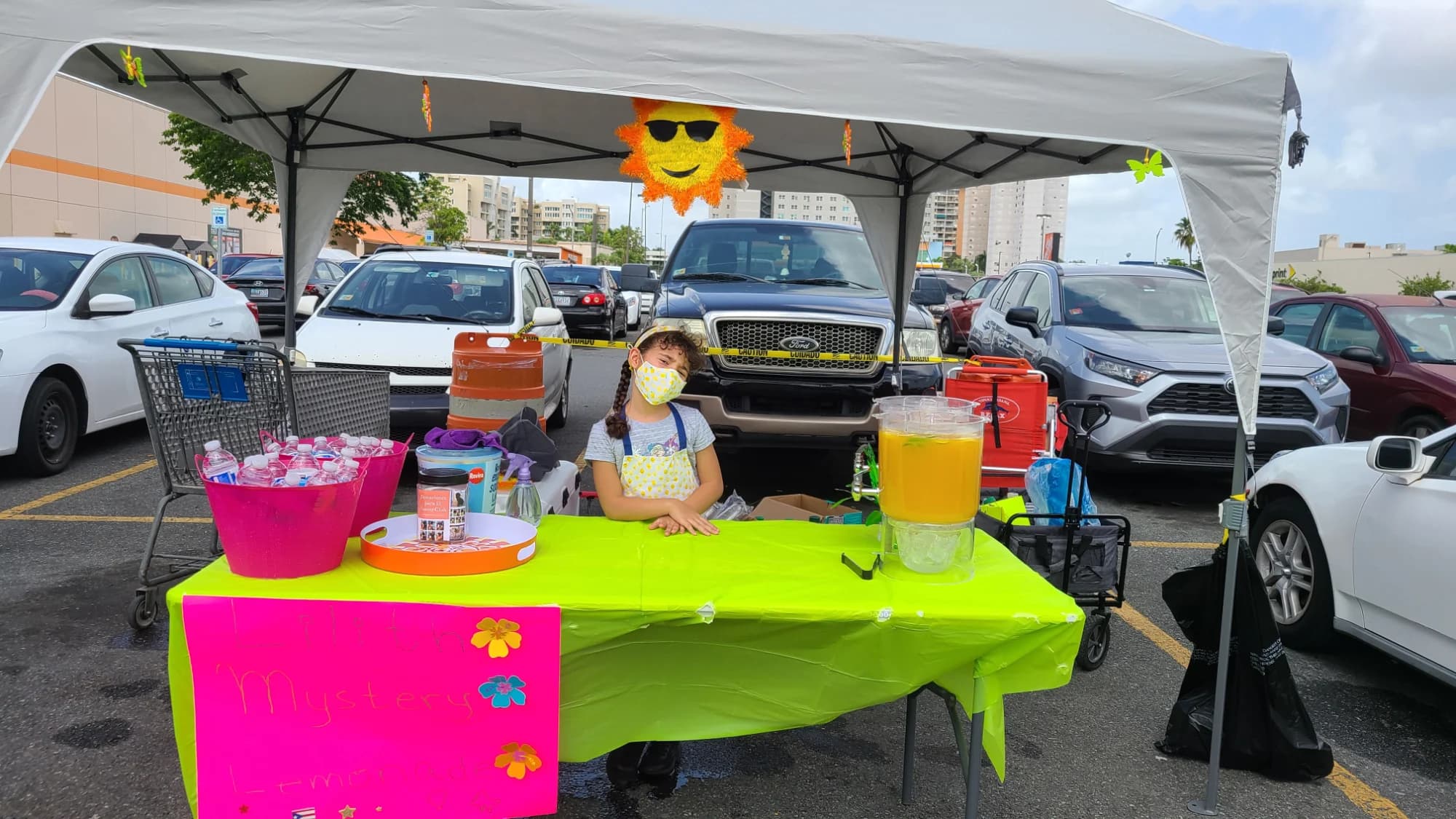 A girl at a bright green and pink lemonade stand in a parking lot