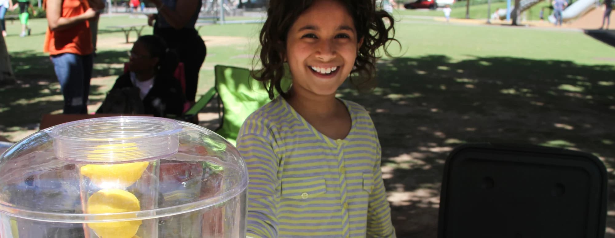 A smiling girl behind a pitcher of lemonade at her stand
